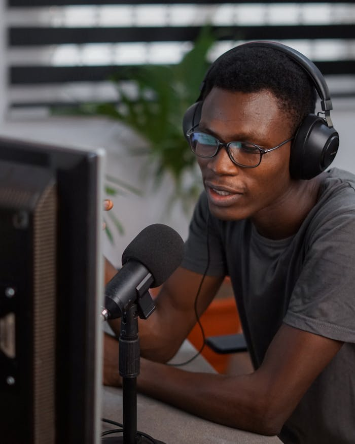 A young man wearing headphones and eyeglasses podcasting indoors with a microphone.